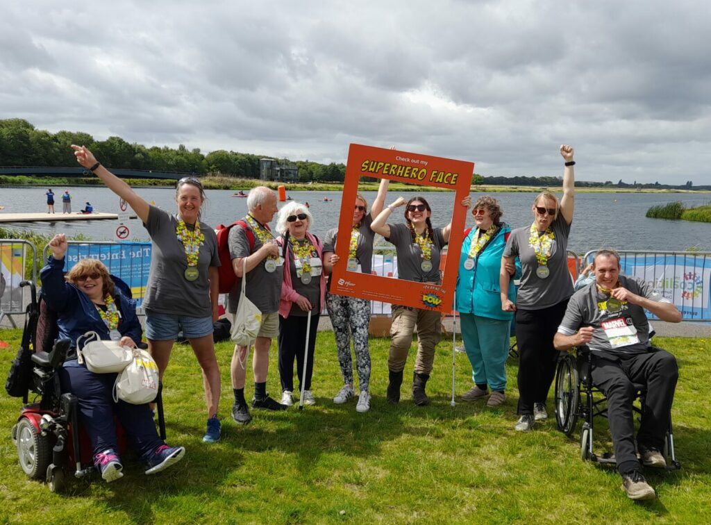 Group photo of the Coalition Superhero Tri participants at the event in 2023 in front of a large lake. All of the participants are wearing a large yellow and silver medal.
