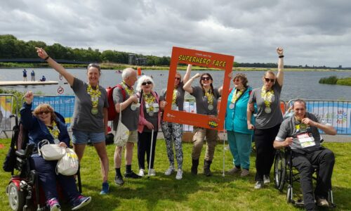 Group photo of the Coalition Superhero Tri participants at the event in 2023 in front of a large lake. All of the participants are wearing a large yellow and silver medal.