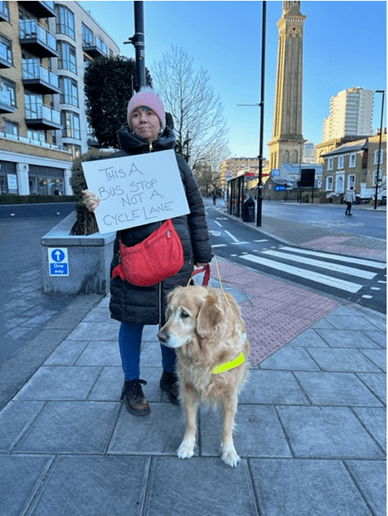 Sarah, from the National Federation of the Blind UK and her guide dog in front of a floating bus stop. Holding a sign saying 'this is a bus stop not a cycle lane'. 